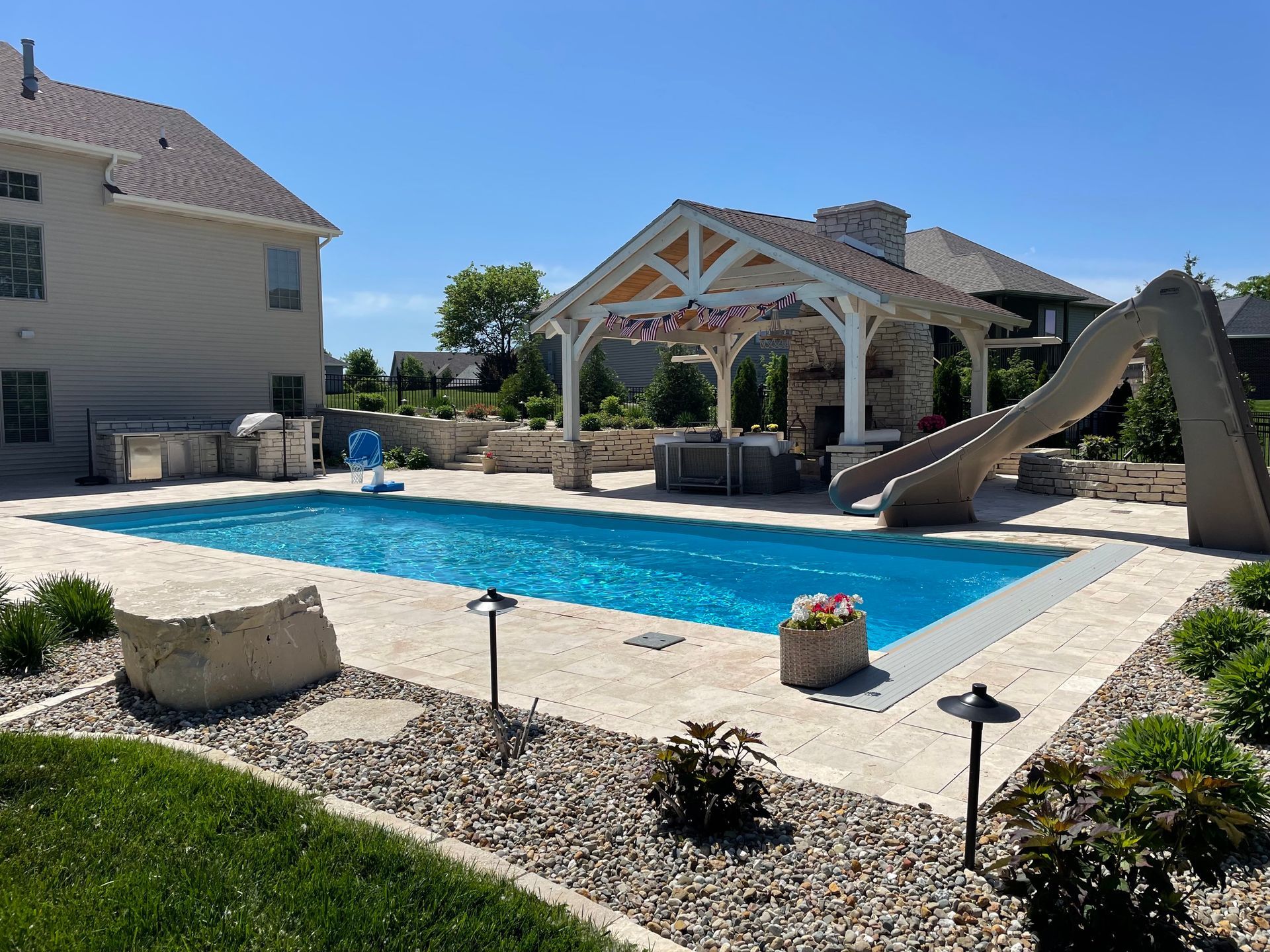 Backyard pool with a slide, outdoor kitchen, and a gazebo on a sunny day.