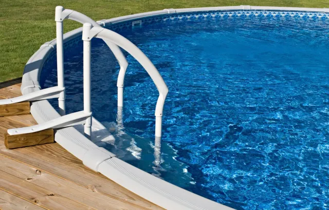 Blue above-ground pool with white ladder on a wood deck, green grass background.