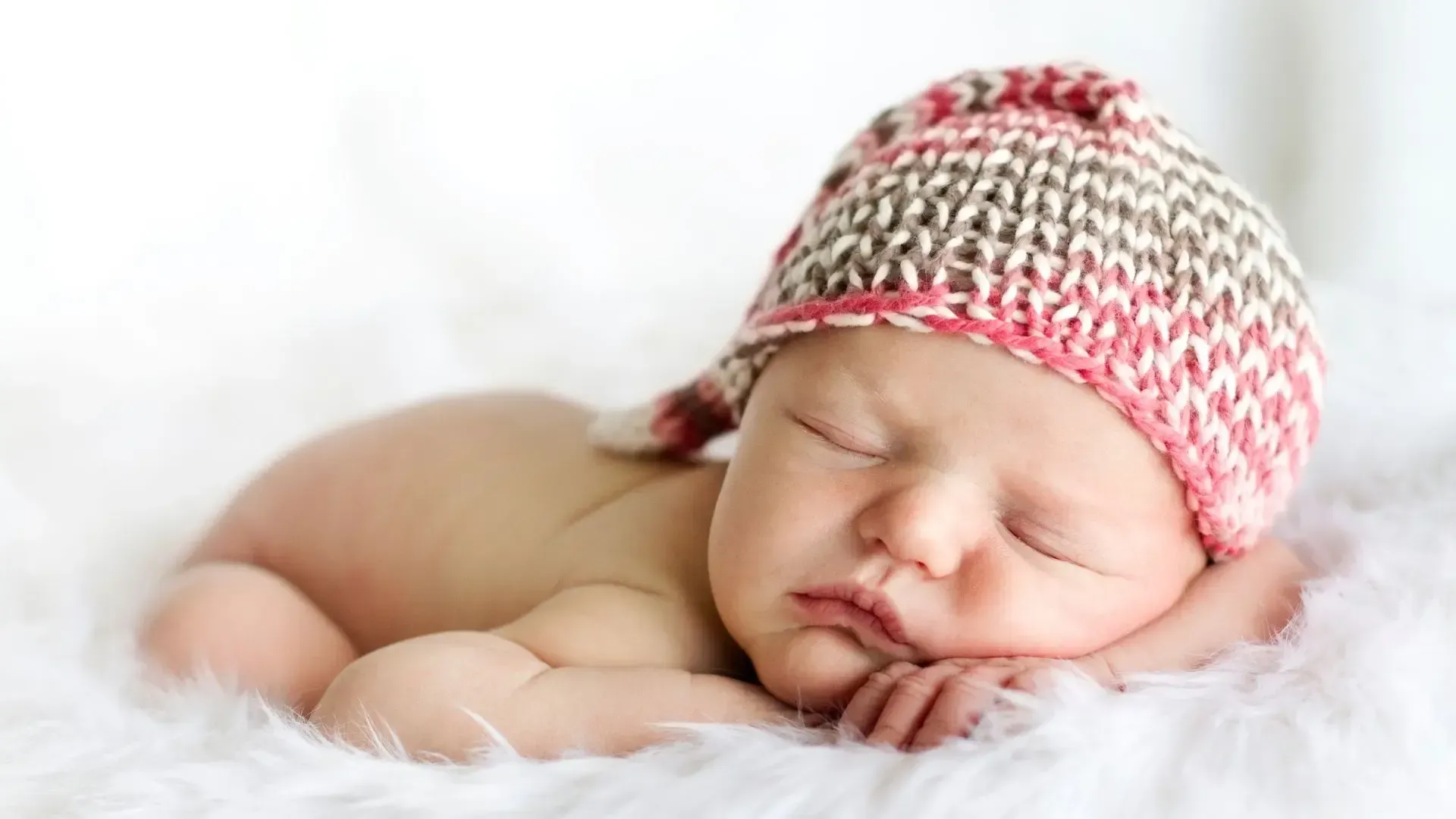 A newborn baby wearing a knitted hat is sleeping on a white blanket.