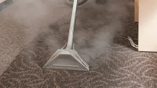 Carpet being steam cleaned with a machine, releasing vapor.