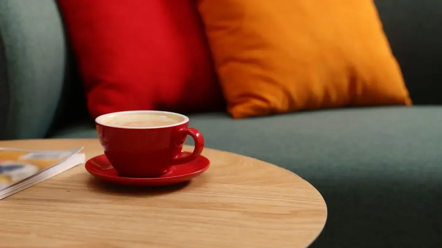 Red coffee cup on wooden table in front of red and orange pillows on a teal couch.