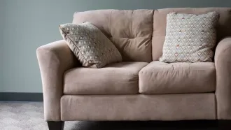 Beige two-seater sofa with two decorative pillows, against a teal wall.
