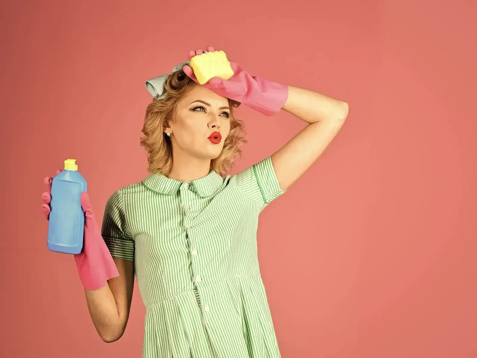 Person in pink gloves and green striped shirt holds cleaning spray and wipes forehead against a pink background