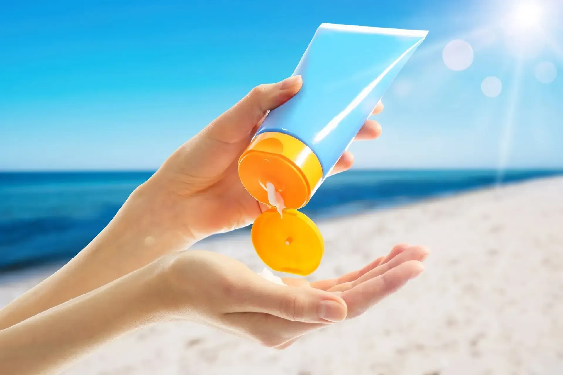 Hands squeezing sunscreen from a blue tube onto a palm at the beach.