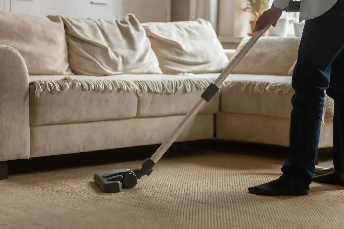 Person vacuuming a beige carpet near a light-colored couch.