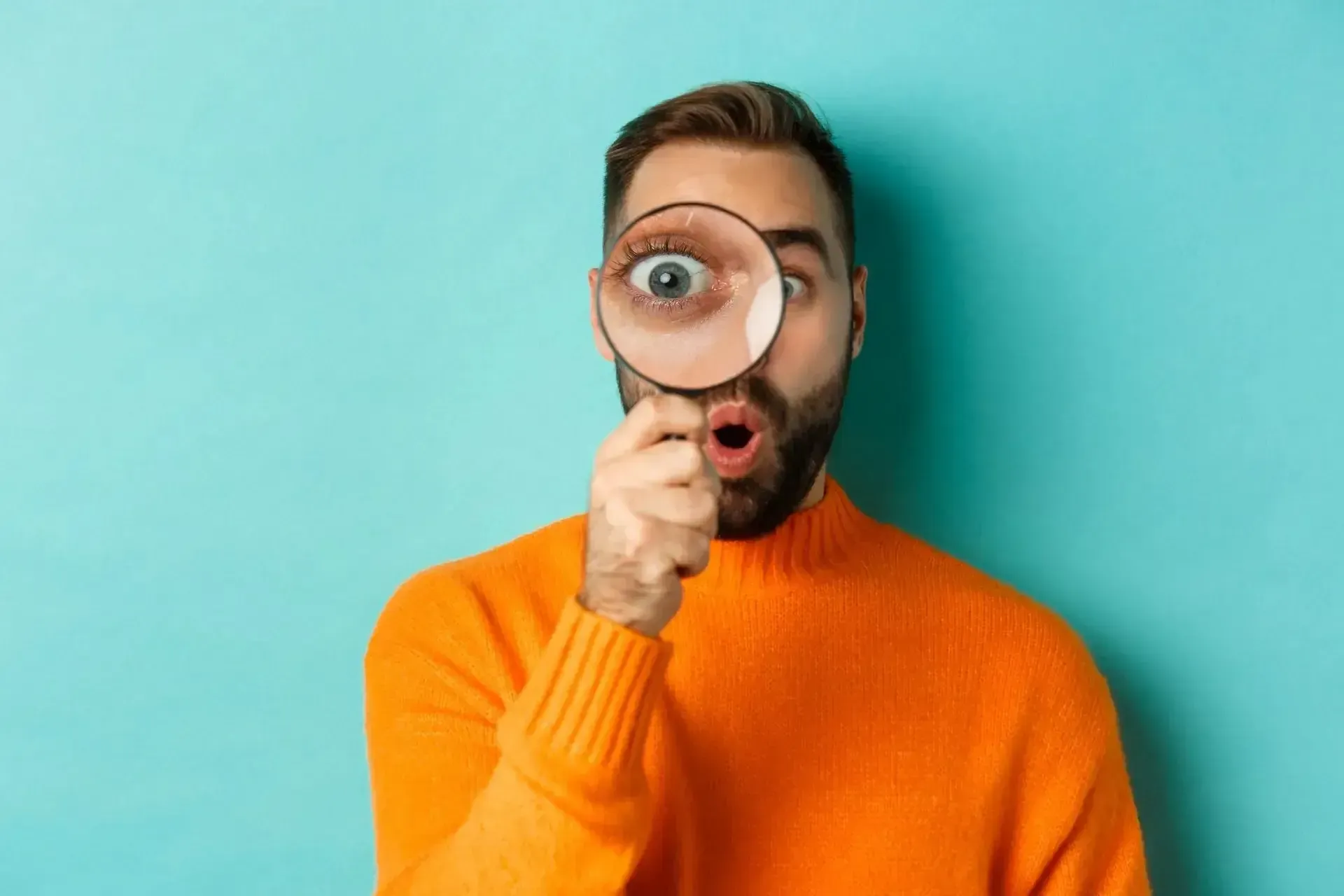 Man with orange sweater uses magnifying glass, eye enlarged; surprised expression. Turquoise background.