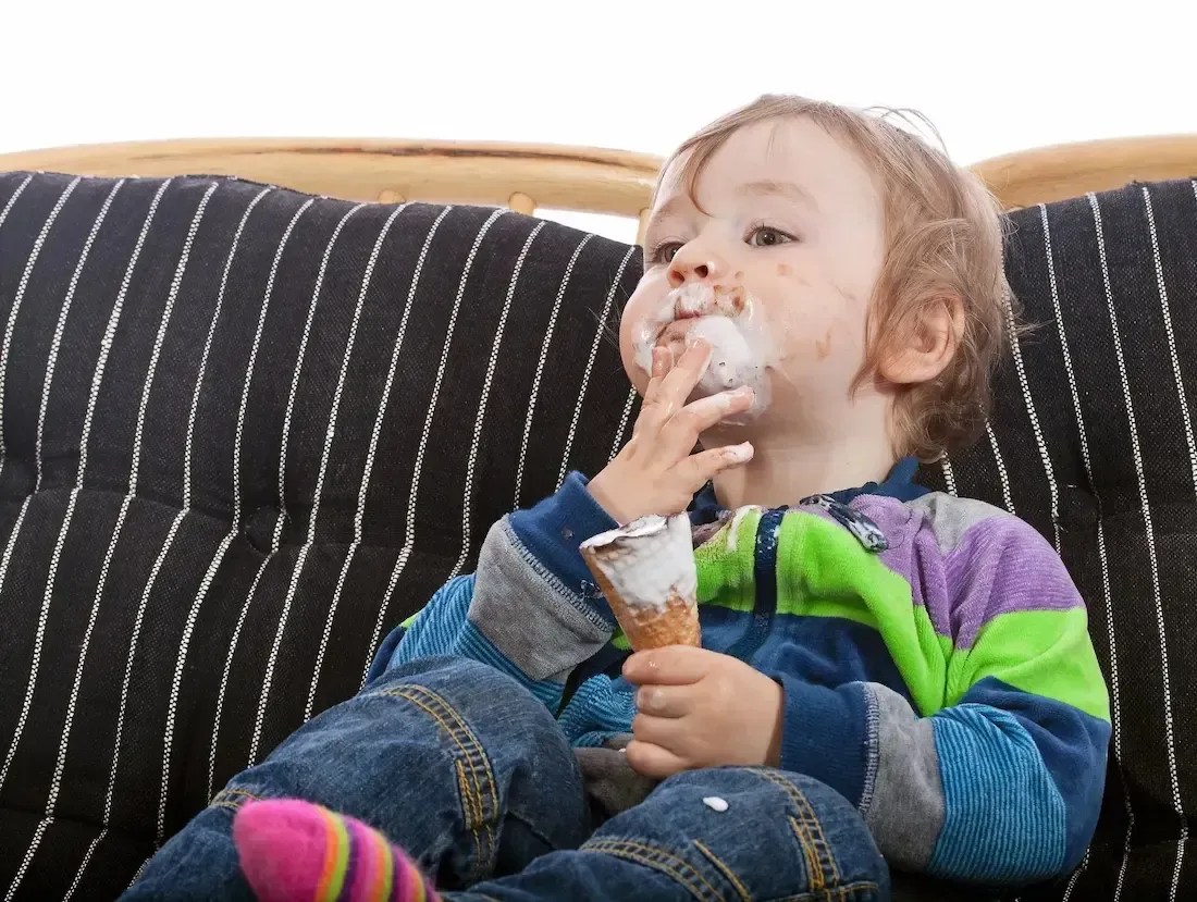 Child eating ice cream cone, face and clothes smeared, seated on striped couch.