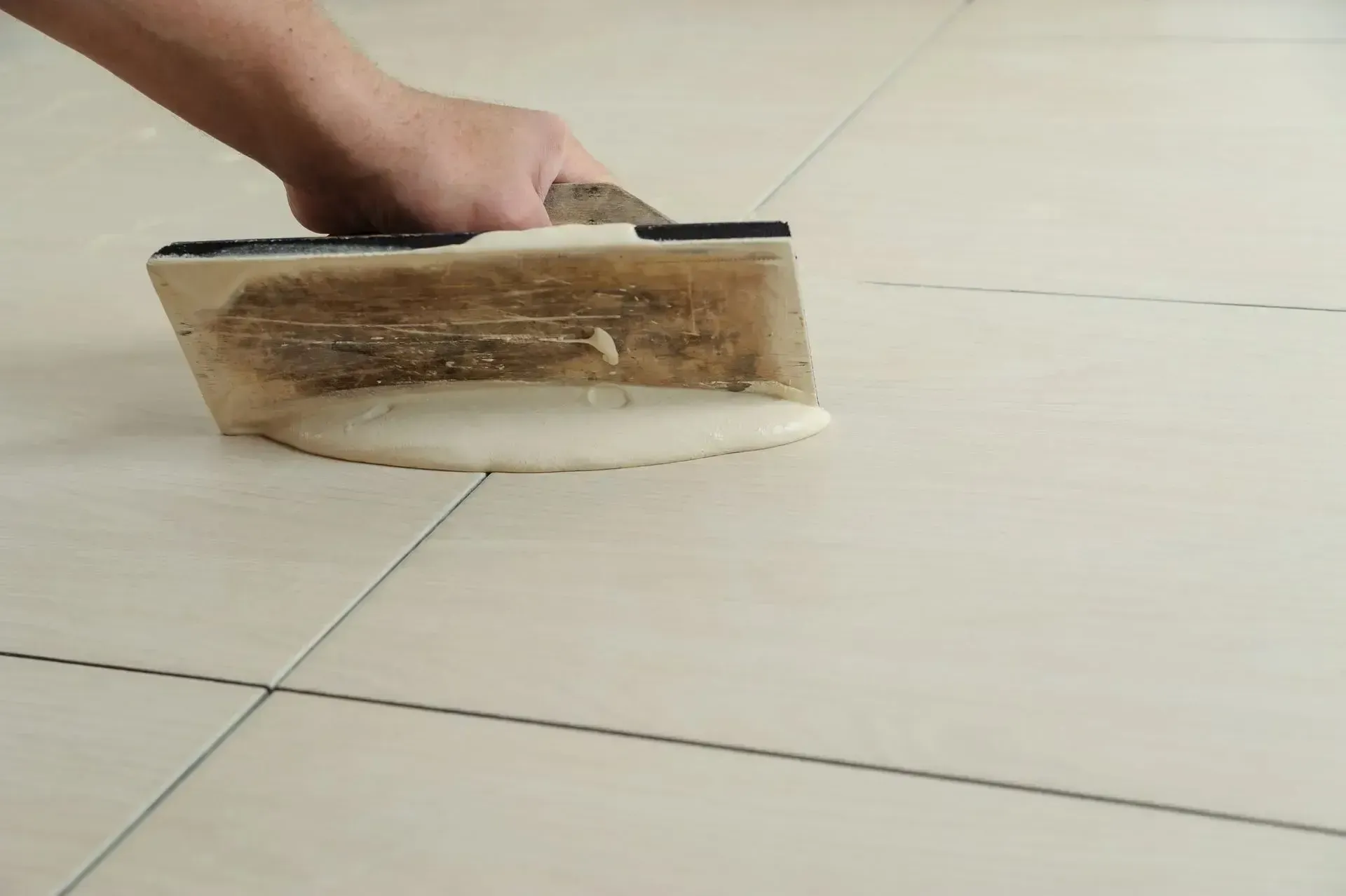 Hand using a trowel to apply grout to floor tiles.