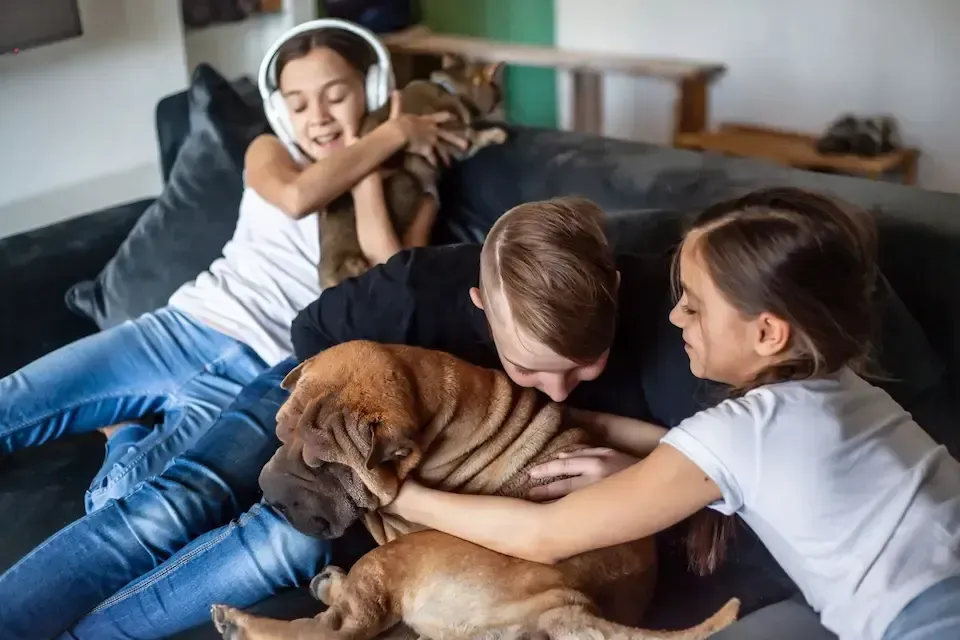 Two children and a woman cuddle a brown dog on a couch; one girl wears headphones and smiles.