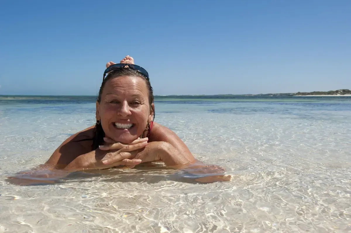 Woman smiling, in shallow, clear ocean water, with hands clasped under chin, sunny day.