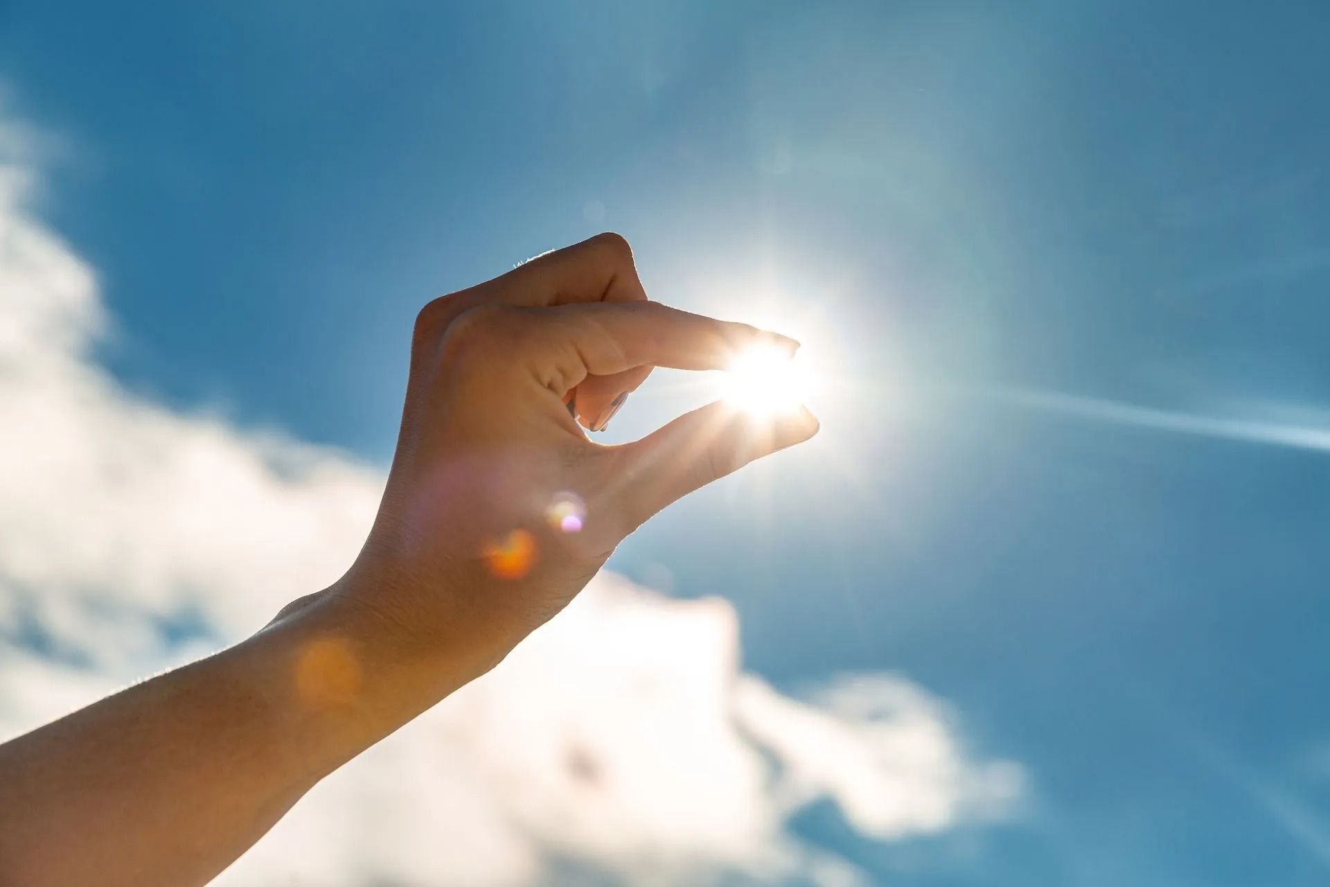 Hand holding up the sun, blue sky with clouds in the background.