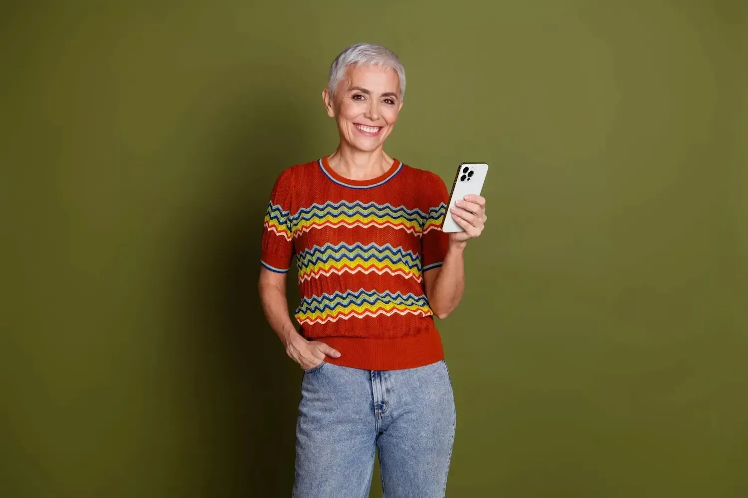 Woman with short gray hair holding a smartphone, wearing a red patterned top and blue jeans, smiling in front of a green backdrop.