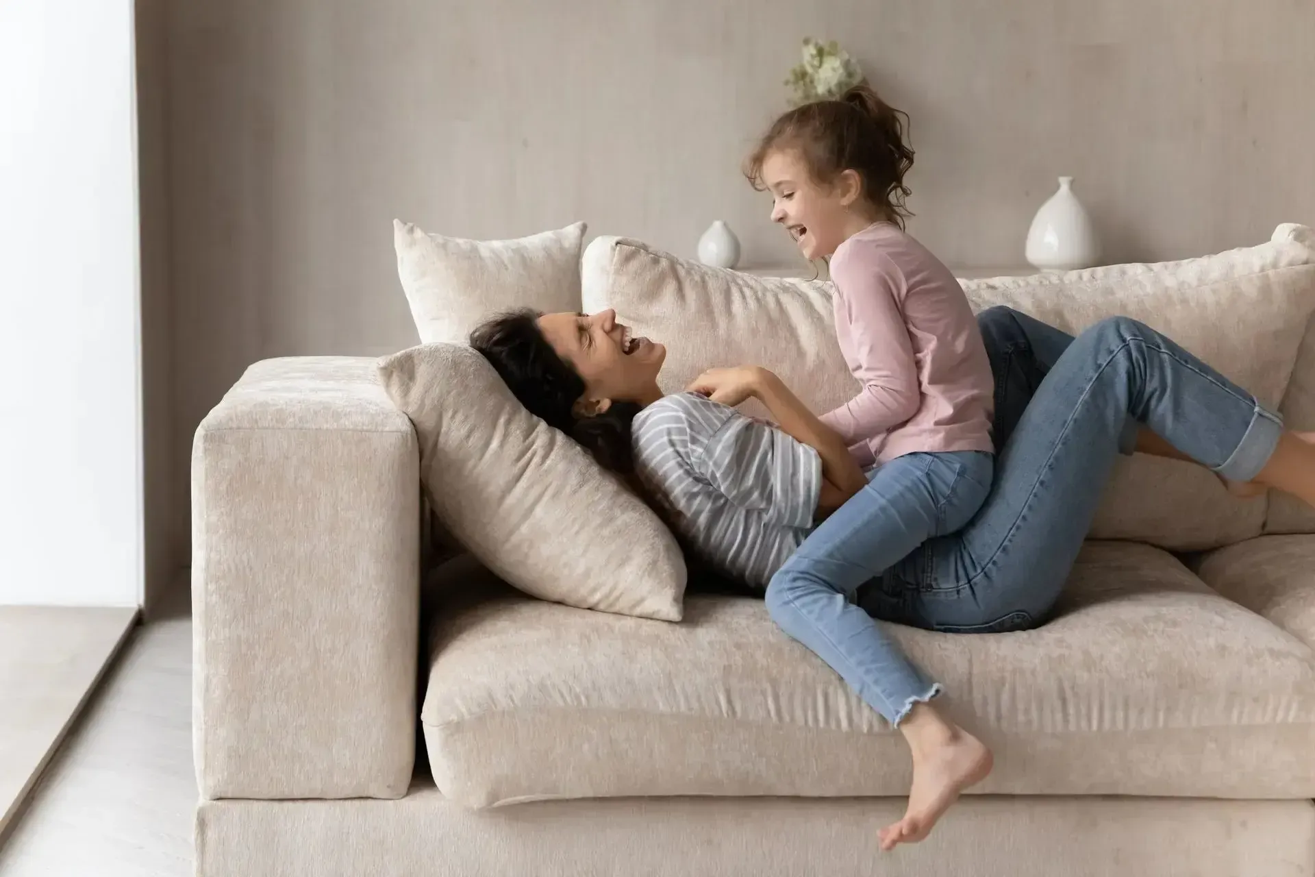 Woman and child laughing, playing on a beige couch indoors.
