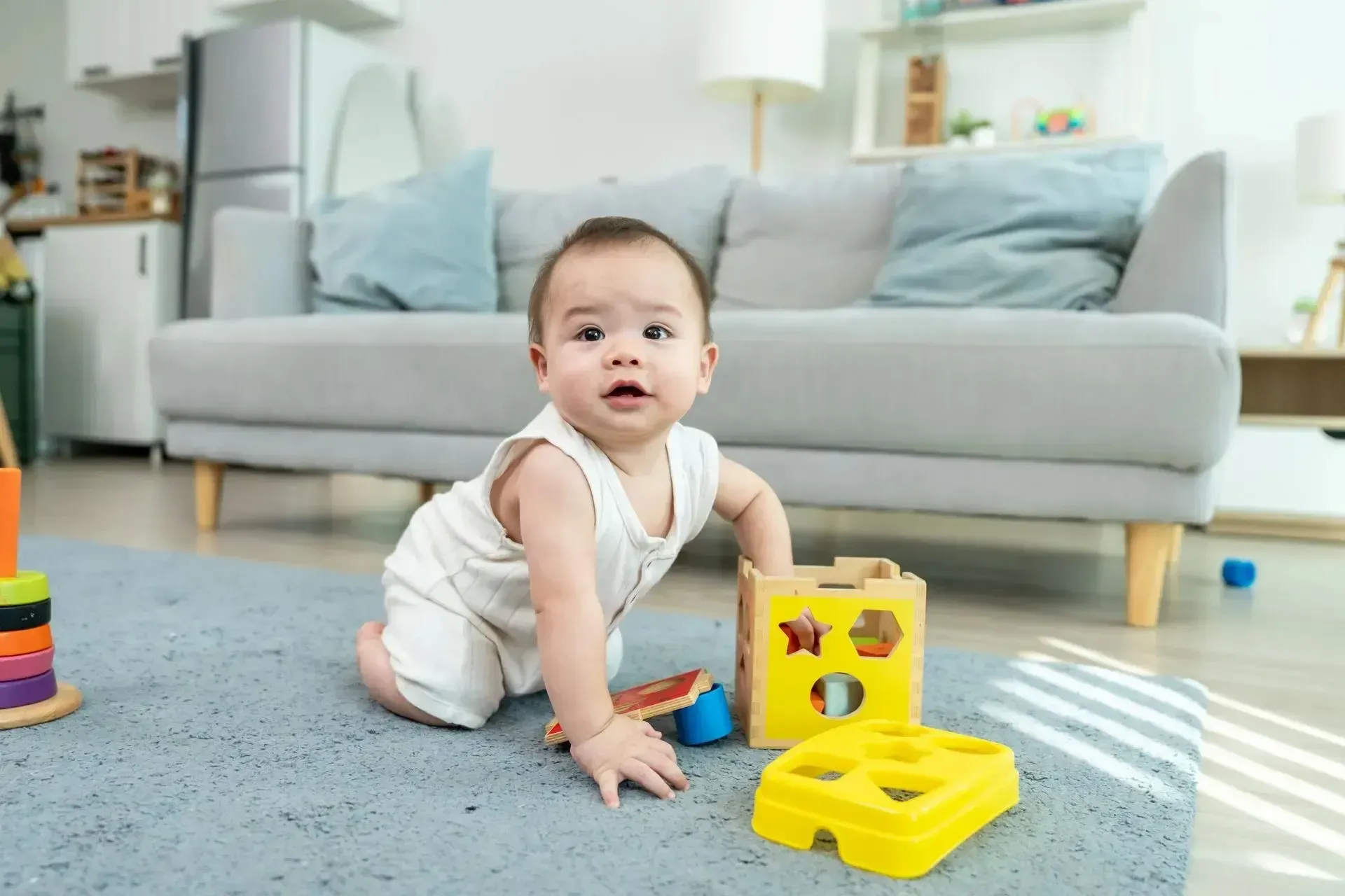 Baby playing on a blue rug, reaching for a yellow shape sorter. Gray couch and living room in background.