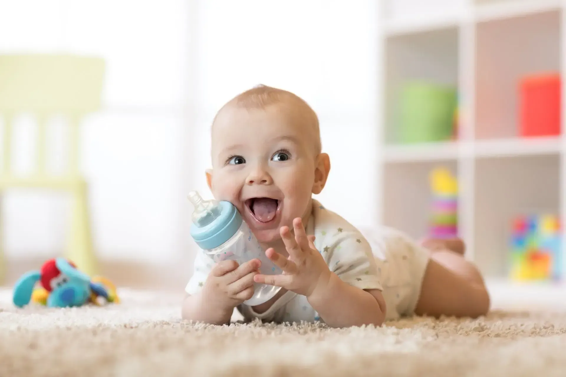 A baby is laying on his stomach on the carpet holding a bottle.