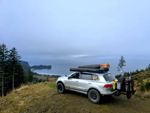 Silver SUV with roof cargo and bike rack overlooking a foggy coastal bay at sunrise