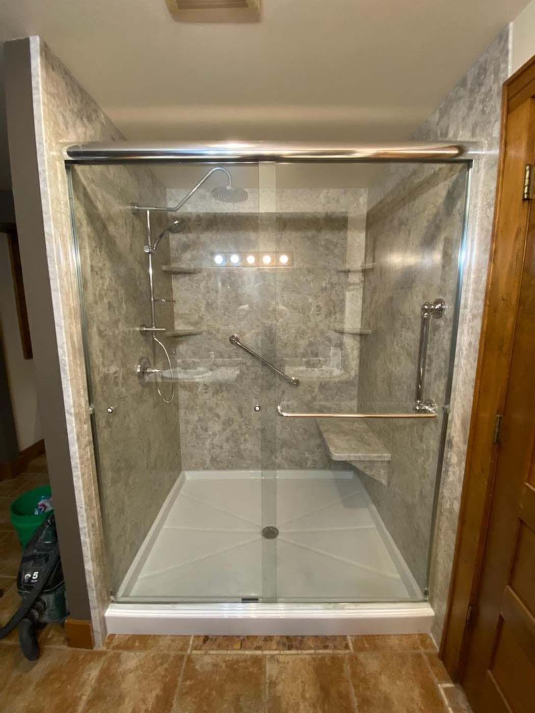 A shower with glass doors, built-in shelves, a seat, and grab bars. The walls and floor are gray.