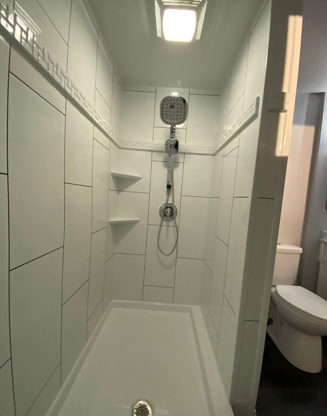 White tiled shower stall with shower head and two shelves. Toilet is visible on the right.