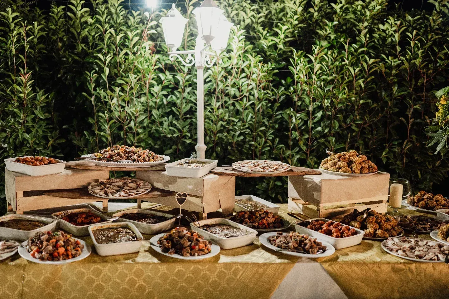 Un tavolo da buffet imbandito con vari piatti, allestito all'aperto sotto un lampione, con uno sfondo verde.