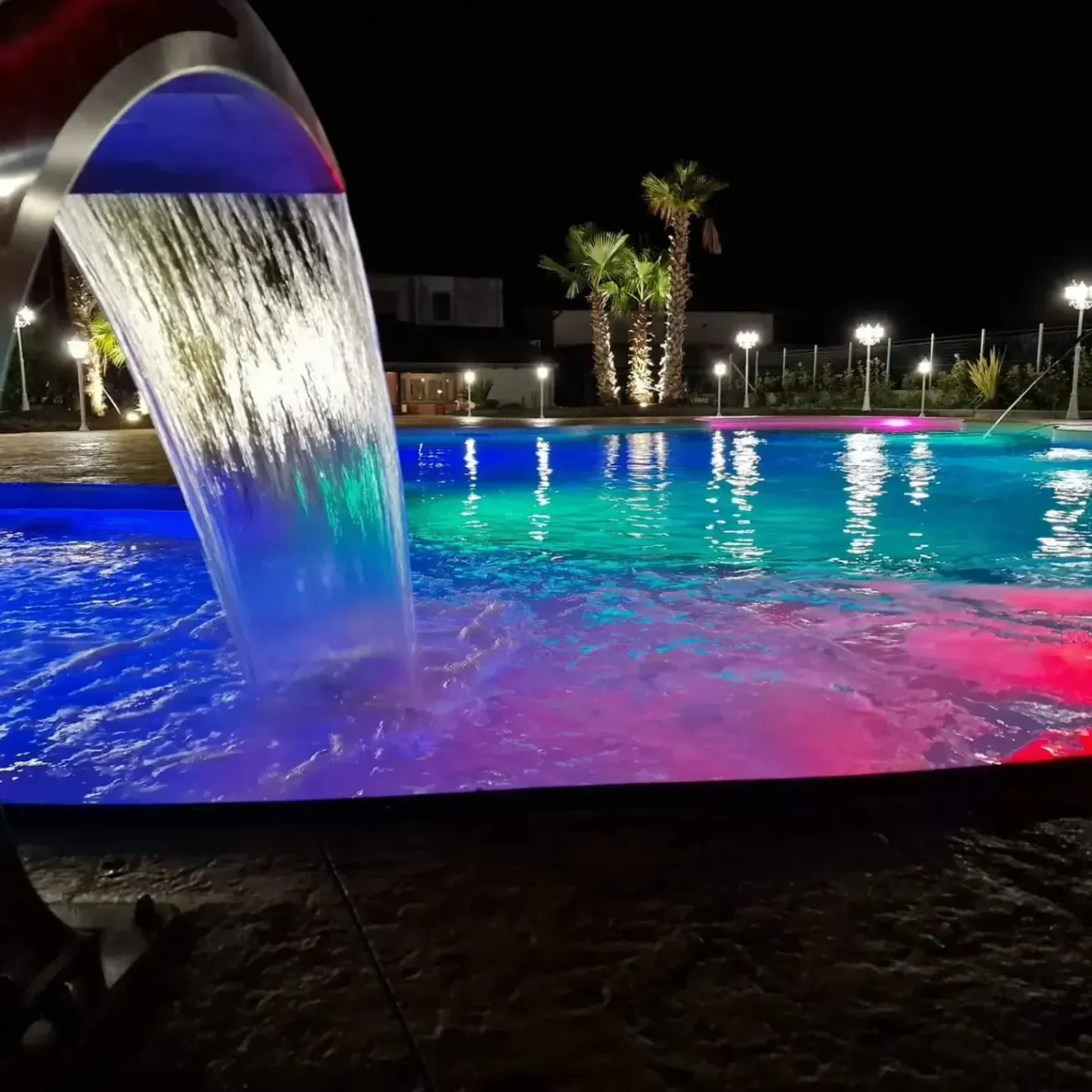 Piscina illuminata di notte con cascata. L'acqua si illumina di blu, verde e rosa. Palme sullo sfondo.