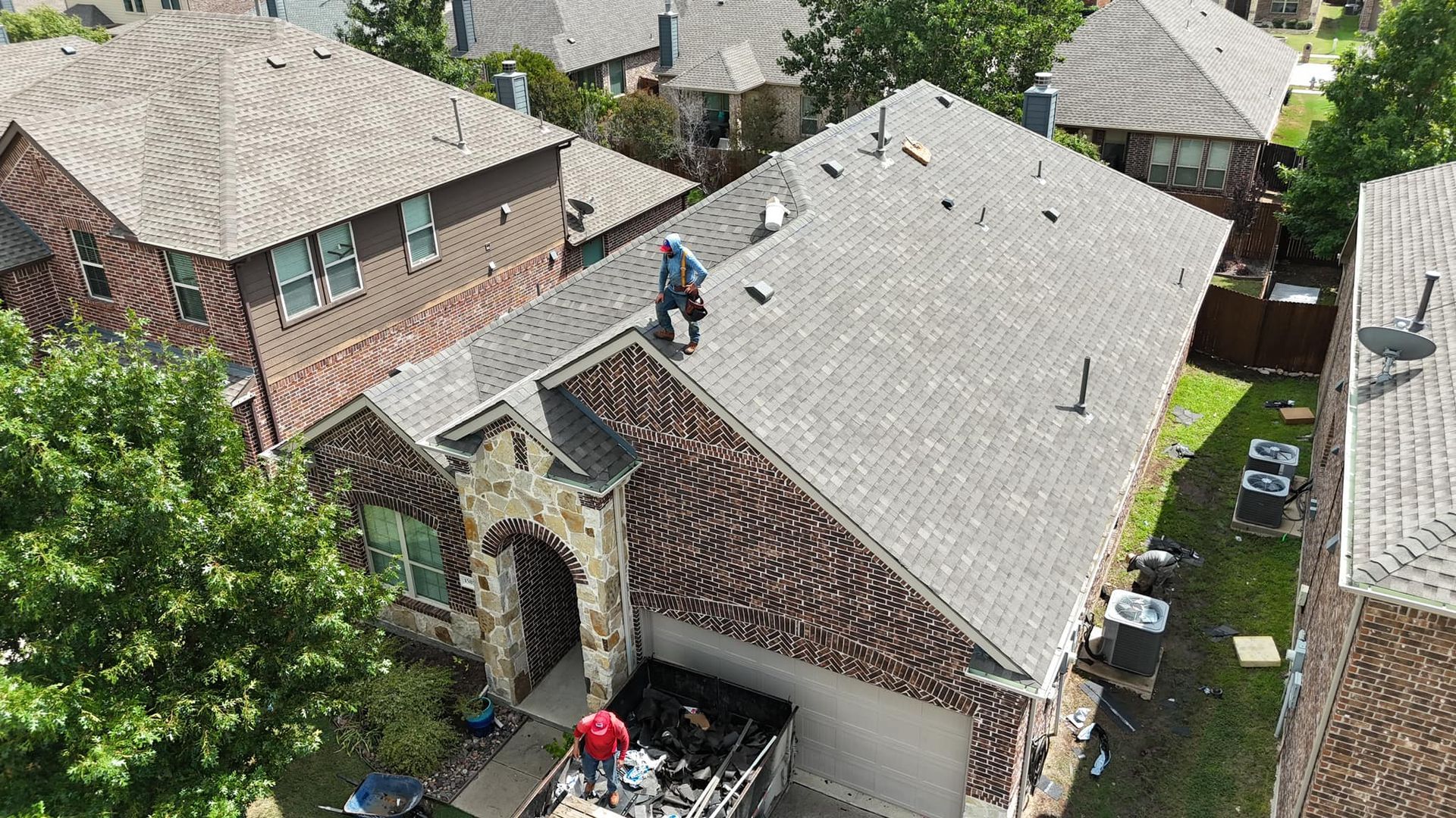 Workers on a rooftop repairing shingles beside a brick house and tall chimney in a neighborhood.