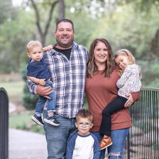 Family of four posing outdoors in casual clothes, smiling with two small children and a fence in the background