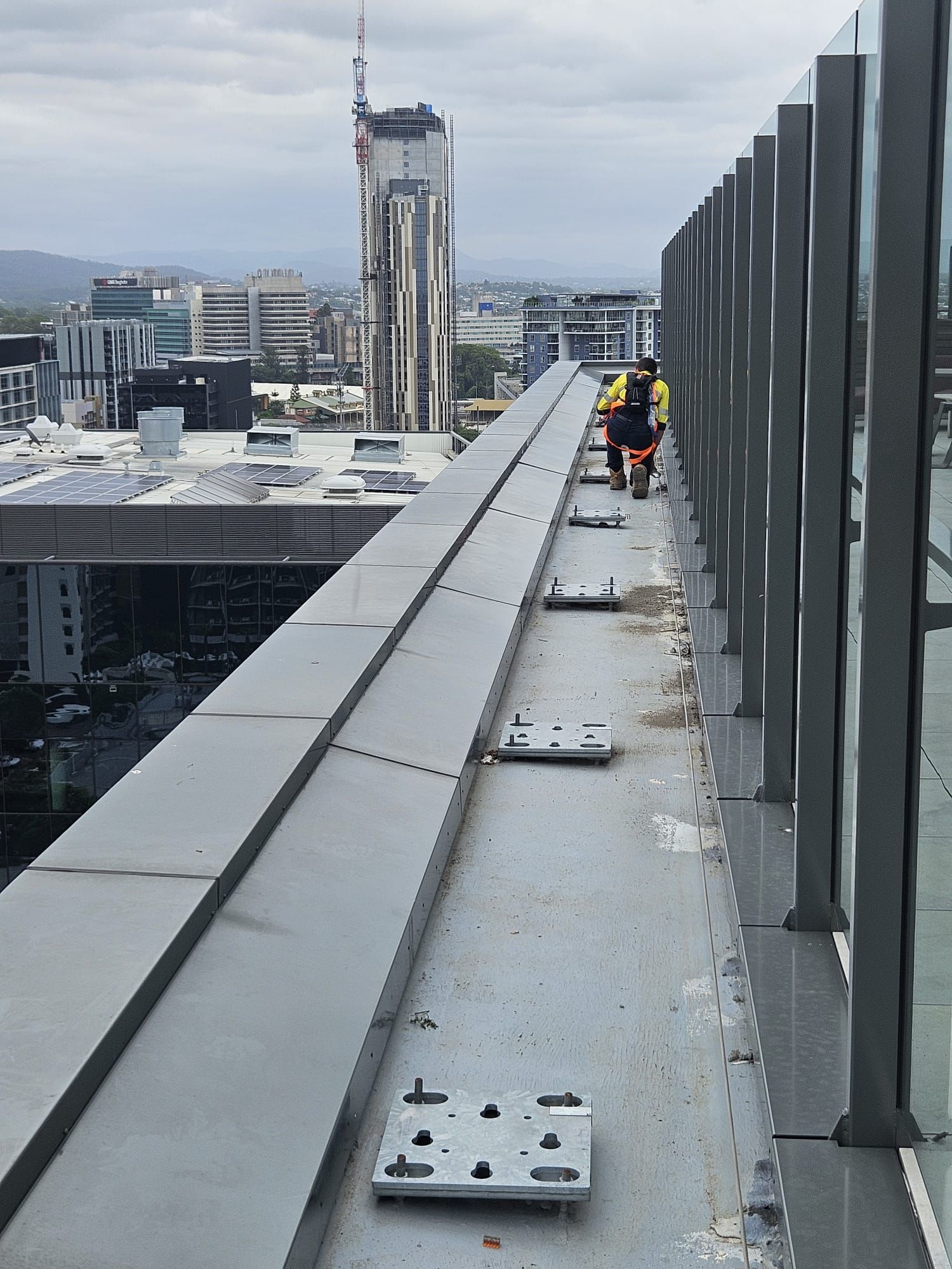 Person working on a rooftop ledge with a city backdrop. Gray building materials and metal fixtures. Overcast day. — Australian Asset Compliance in Gympie, QLD