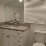 Bathroom with white cabinets, granite countertop, and a toilet. Mirror and window with blinds visible.