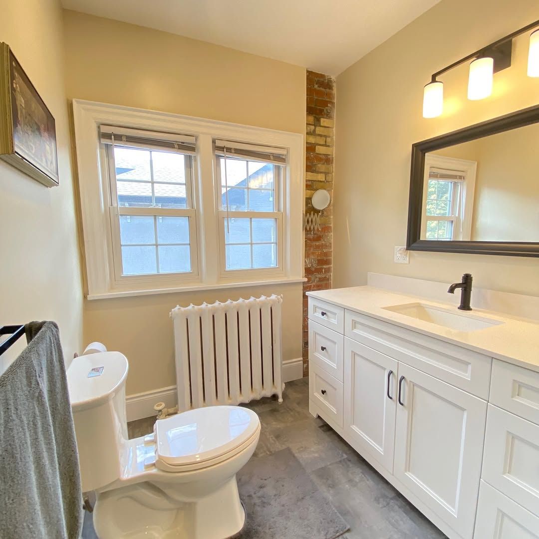Bathroom with white vanity, toilet, and radiator. Window offers natural light. Exposed brick wall.