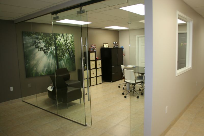 Glass-walled office with a painting, chair, round table, and a hallway with a window.