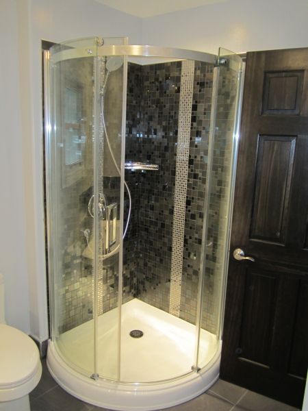 Curved glass shower stall with dark mosaic tile backdrop next to a dark wood door.