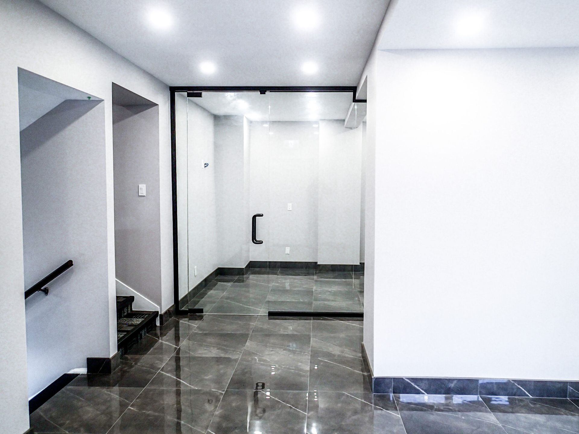 Empty, modern hallway with glass door entrance, gray tile flooring, and staircase on the left.
