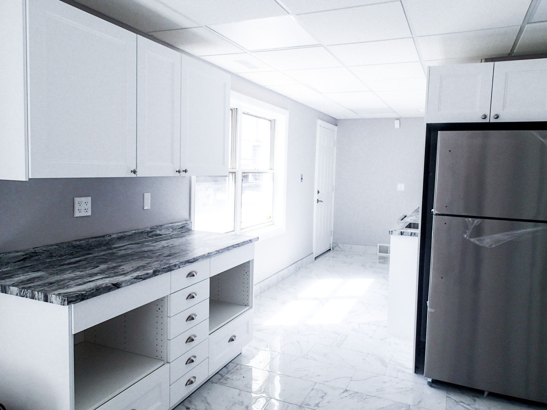 White kitchen with stainless steel refrigerator, gray countertops, and white cabinets.