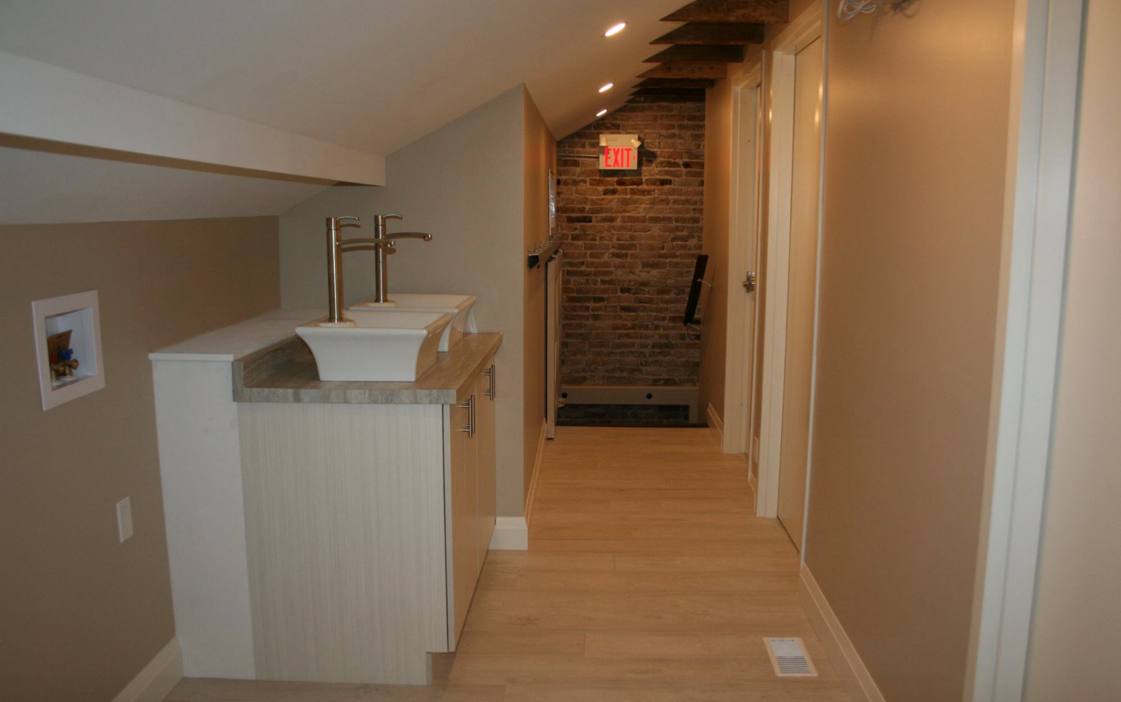Narrow bathroom with a double vanity, wooden floors, and exposed brick wall.