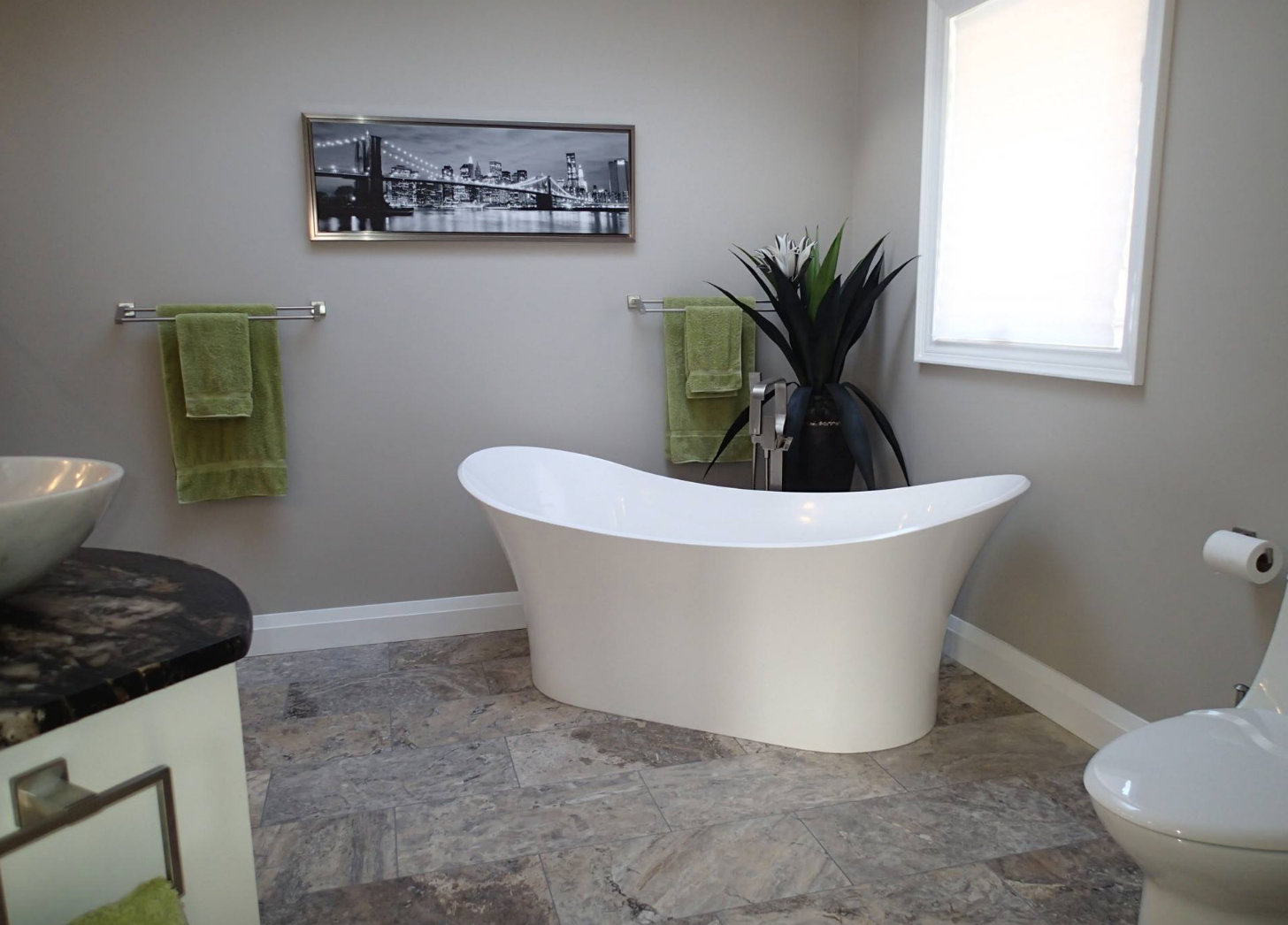 Bathroom with a white tub, green towels, a plant, a window, and a toilet; walls are gray.