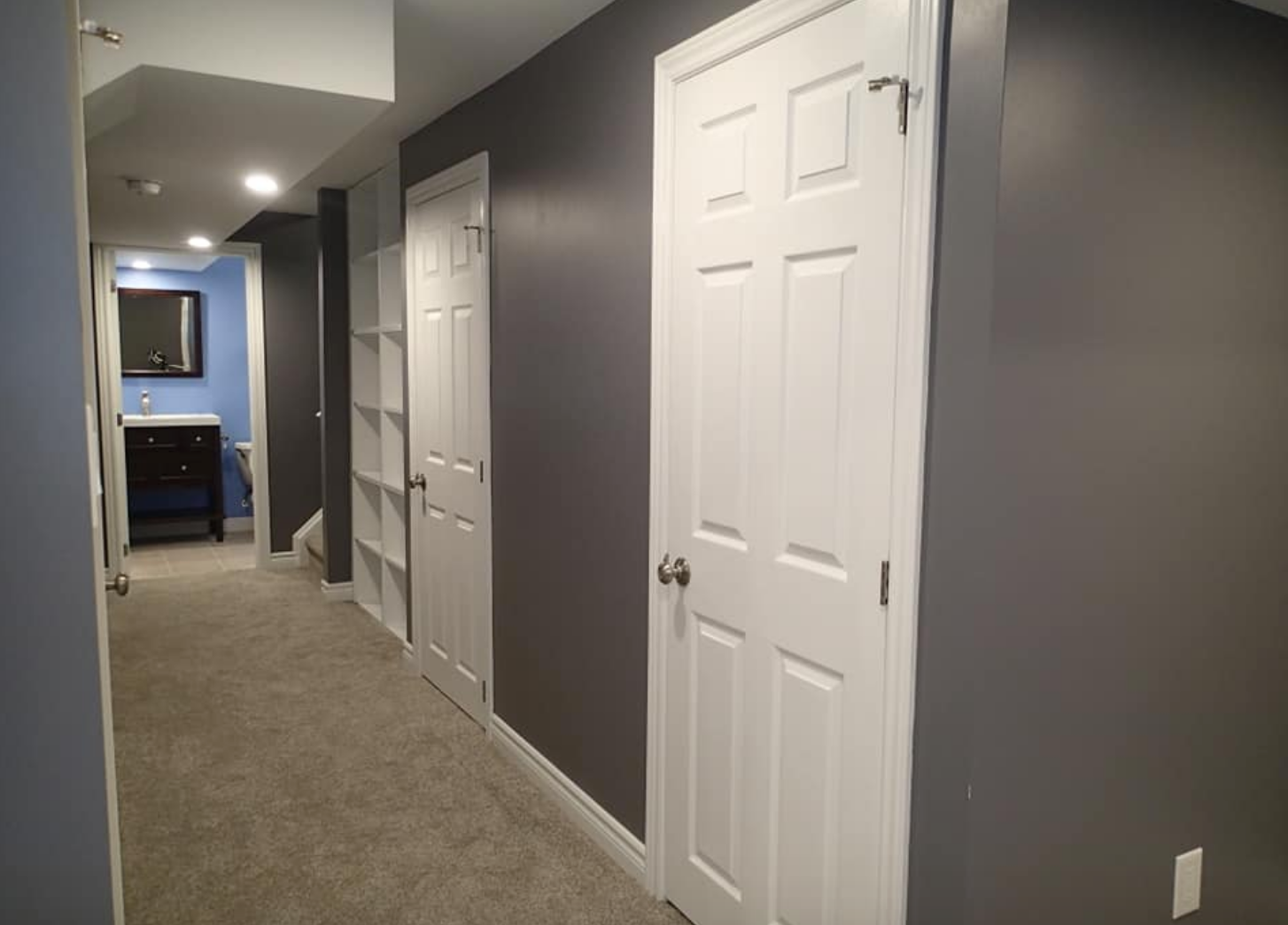 Long hallway with gray walls, white doors, and carpet flooring. A blue room with a sink is at the end.