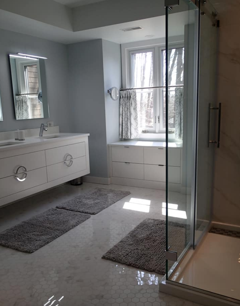 Modern bathroom with a white vanity, glass shower, and natural light from a window. Gray rugs on white tiled floor.