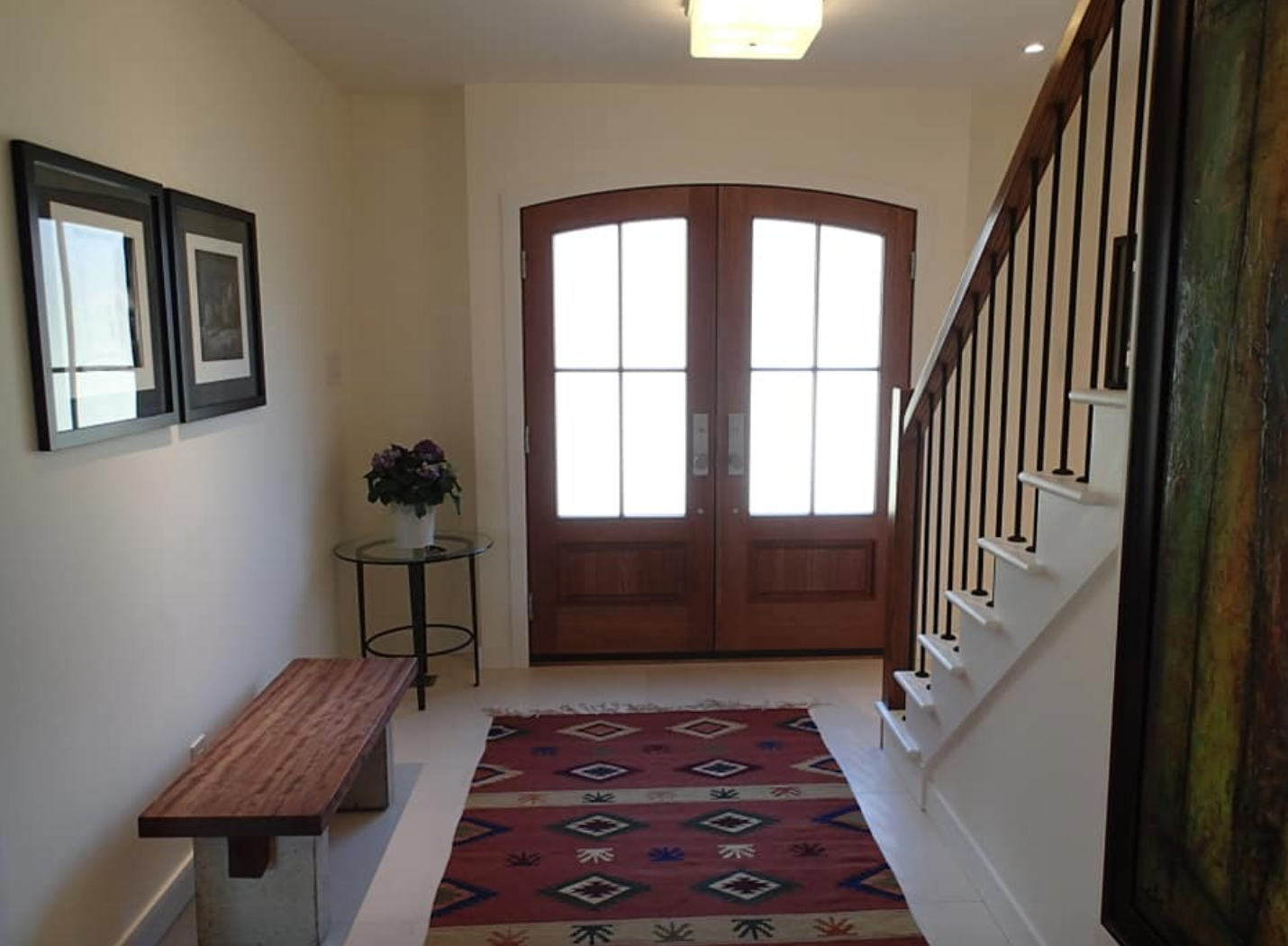 Entryway with wooden double doors, stairs, and a patterned rug. A bench and artwork decorate the space.