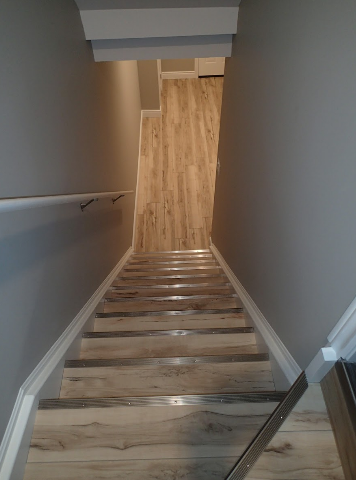 Wooden staircase leading down with gray walls, handrail, and wood-look flooring at the top.
