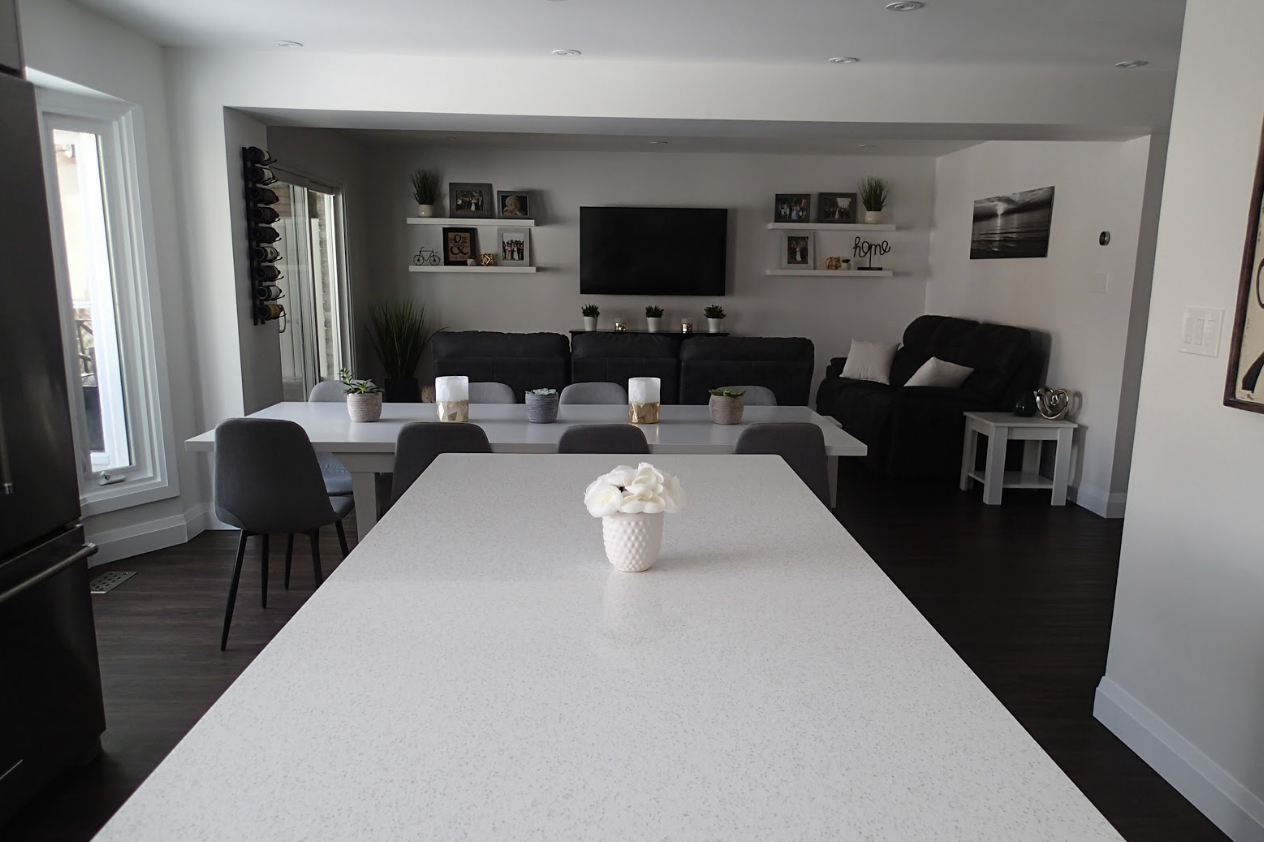 Spacious dining room with a long white table, dark chairs, and a view into the living room with a TV and sofa.