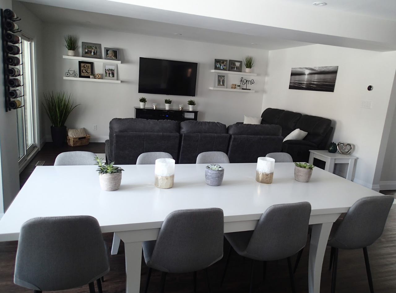 A dining room with a white table, gray chairs, and a sectional sofa. Decorations include plants and framed art.
