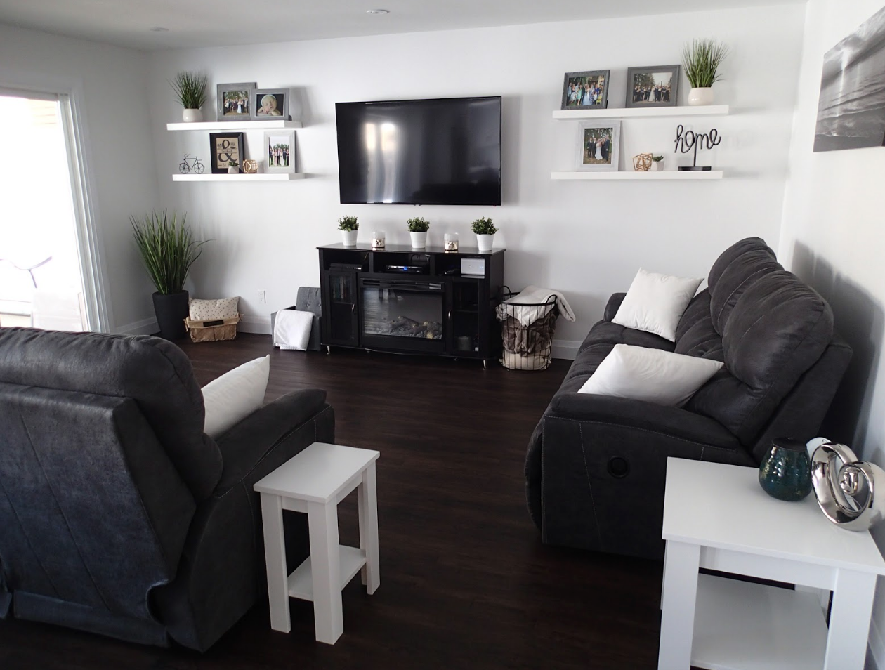 Living room with dark recliner sofas, white side tables, TV, and shelves with decor.