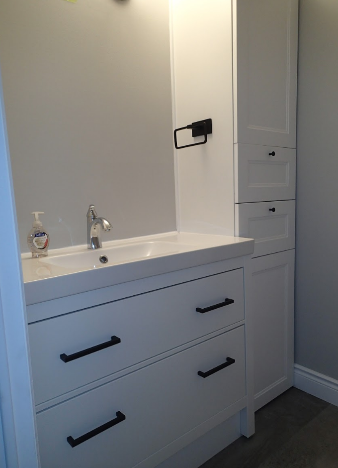 White bathroom vanity with black hardware, beside a tall cabinet, against a light grey wall.