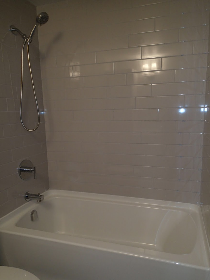 Bathroom with white tub, gray tiled walls, and chrome shower fixtures.