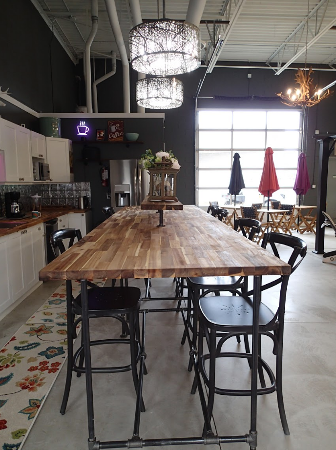 A long wooden table with bar stools in a kitchen area. Overhead lights and open garage door.
