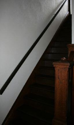 Black handrail along a white wall, leading up dark wooden stairs, with a brown wooden post visible.