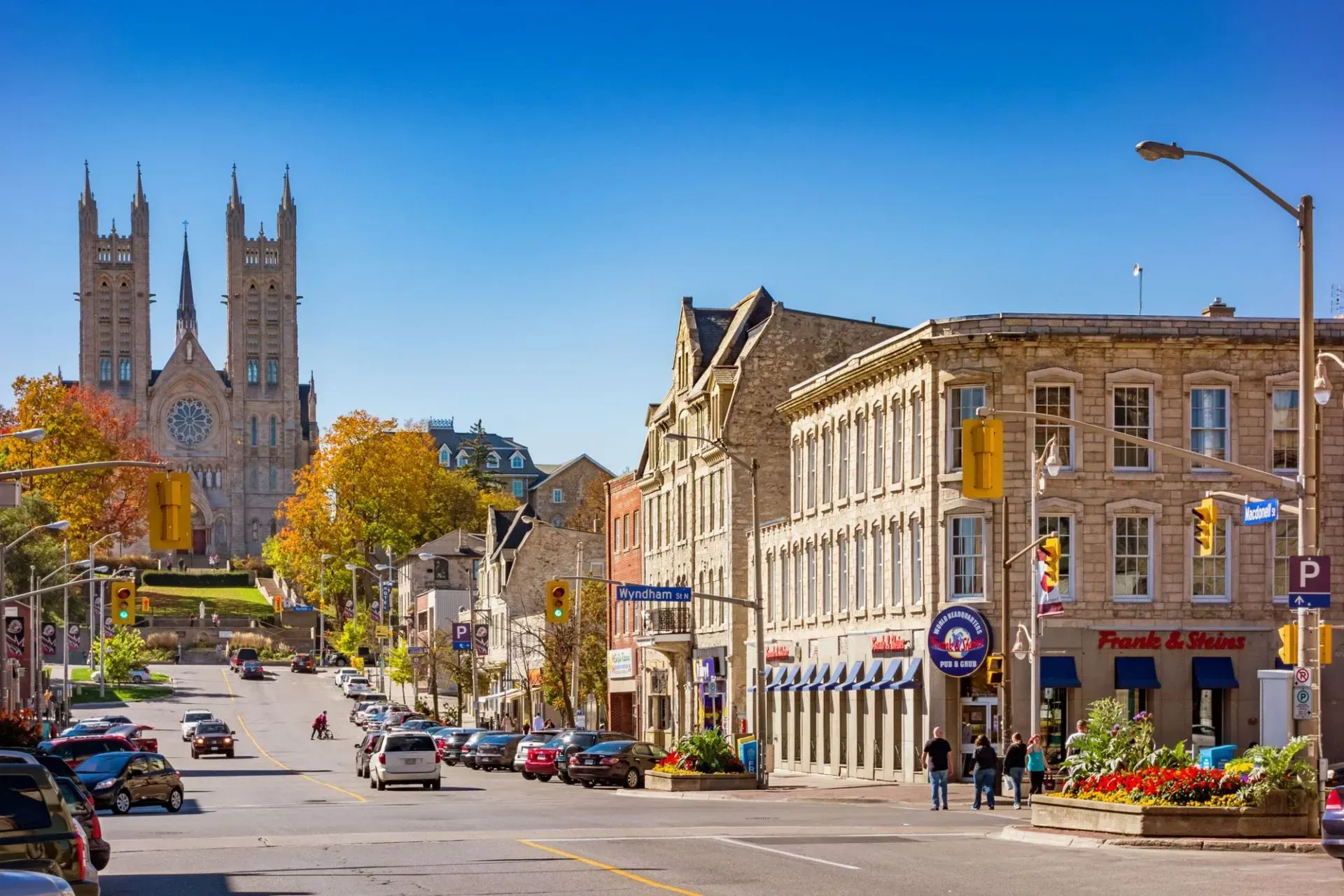 Street view with stone buildings, church towers in background, cars, pedestrians, blue sky.