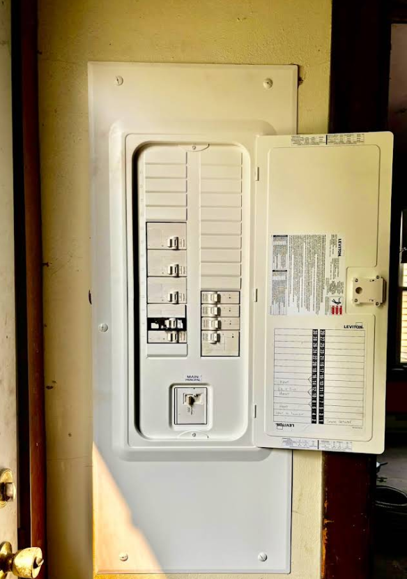 Open electrical circuit breaker panel with white door on a beige wall.