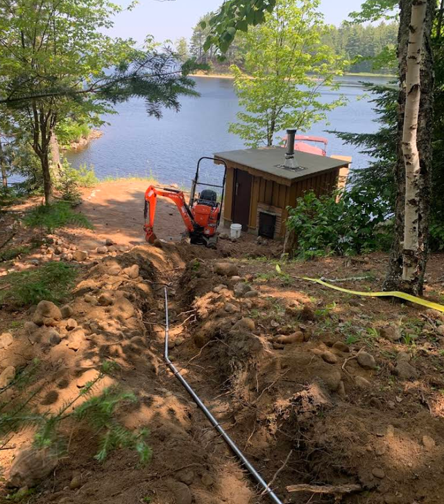 Excavator on a muddy lakeside slope beside a small wooden shed and trees under bright daylight