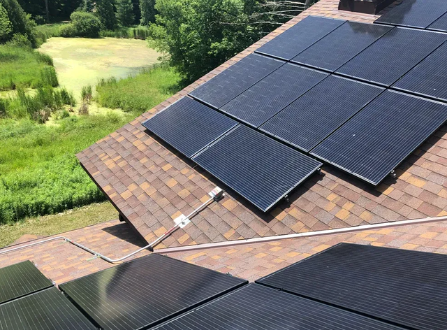 Solar panels on a brown shingled roof beside a green landscape and pond